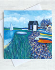 A cute little blck hut sits on the shore alongside the river with a sailing boat passing by. Brightly coloured boats are pile up alongside a pebbled path.