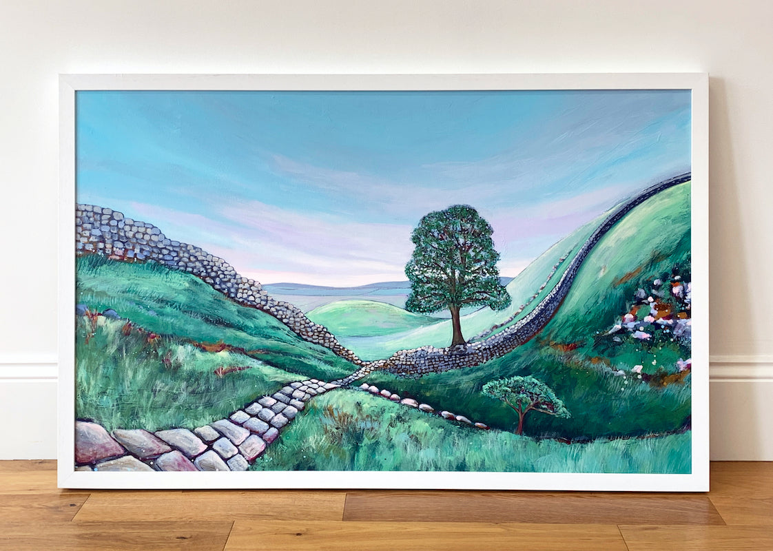 A large painting in a white frame standing on a wooden floor. The painting features the famous Sycamore Gap tree in the Northumberland countryside. The landscape is lush green with a subtle pink in the sky. There  is a cobbled path leading down the hill towards the tree in the dip in the landscape. Hadrians wall perches along the ridge of the hills. 