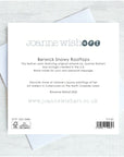 The back of a Joanne Wishart 'Berwick Snowy Rooftops' greeting card, showing printed text and barcode details, displayed on top of white envelopes on a light gray textured background.