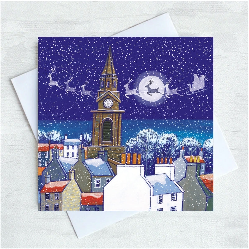 A square Christmas greeting card featuring a snowy night in Berwick village scene with  tower, rooftops, sleighs, and Santa in the sky, shown with a white envelope on a light textured background.