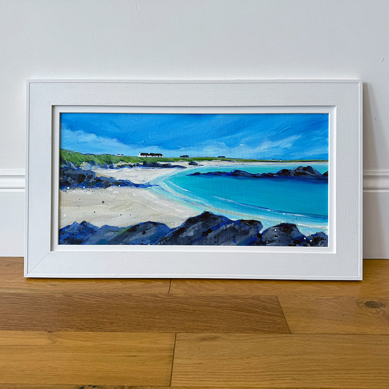 A framed landscape painting of a coastal beach with turquoise water, white sand, dark rocky foreground, and a blue sky, displayed against a white wall on a wooden floor.