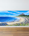 Framed painting of a coastal scene with a castle, beach, and flowers on a wooden floor.