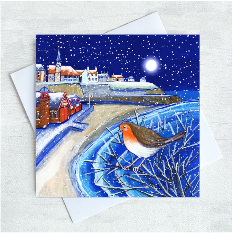 A square Christmas greeting card featuring a winter Cullercoats Bay scene with snow, a starry night sky, and a robin perched in the foreground, shown with a white envelope on a light textured background.