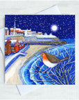 A square Christmas greeting card featuring a winter Cullercoats Bay scene with snow, a starry night sky, and a robin perched in the foreground, shown with a white envelope on a light textured background.