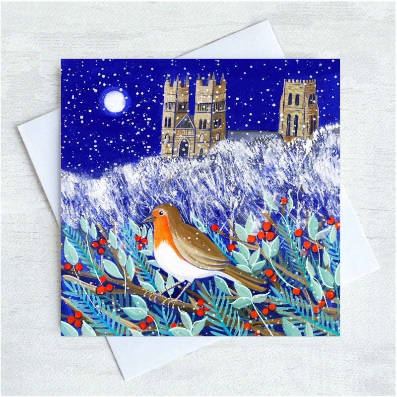 A square greeting card featuring a winter night scene with a robin bird in the foreground, holly branches with red berries, and cathedral towers under a snowy blue sky, shown with a white envelope on a light textured background.