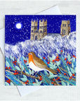 A square greeting card featuring a winter night scene with a robin bird in the foreground, holly branches with red berries, and cathedral towers under a snowy blue sky, shown with a white envelope on a light textured background.