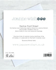 The back of a Joanne Wishart 'Festive Front Street' card, showing printed text, barcode, and recycling symbol, resting on top of a white envelope against a light gray textured background.