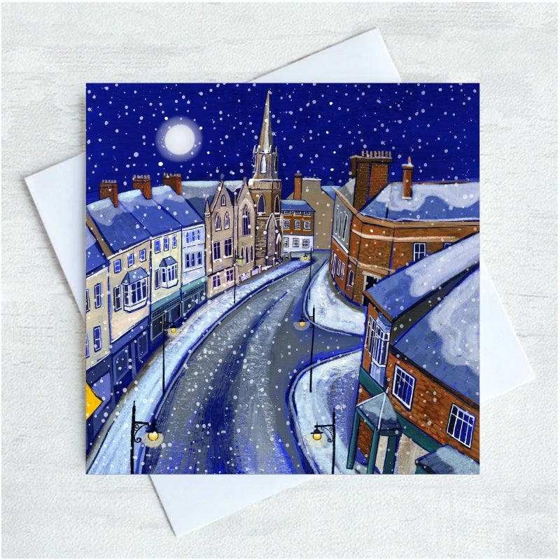A square art print or card showing a snowy night tynemouth front street scene with buildings, a church spire, and a full moon, displayed over a white envelope on a light textured background.