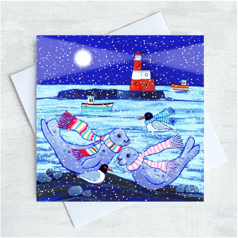 A square illustrated greeting card featuring seals, penguins, boats, and a lighthouse in a snowy night scene in the Farnes shown with a white envelope on a light textured background.