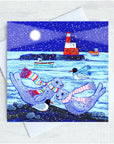 A square illustrated greeting card featuring seals, penguins, boats, and a lighthouse in a snowy night scene in the Farnes shown with a white envelope on a light textured background.