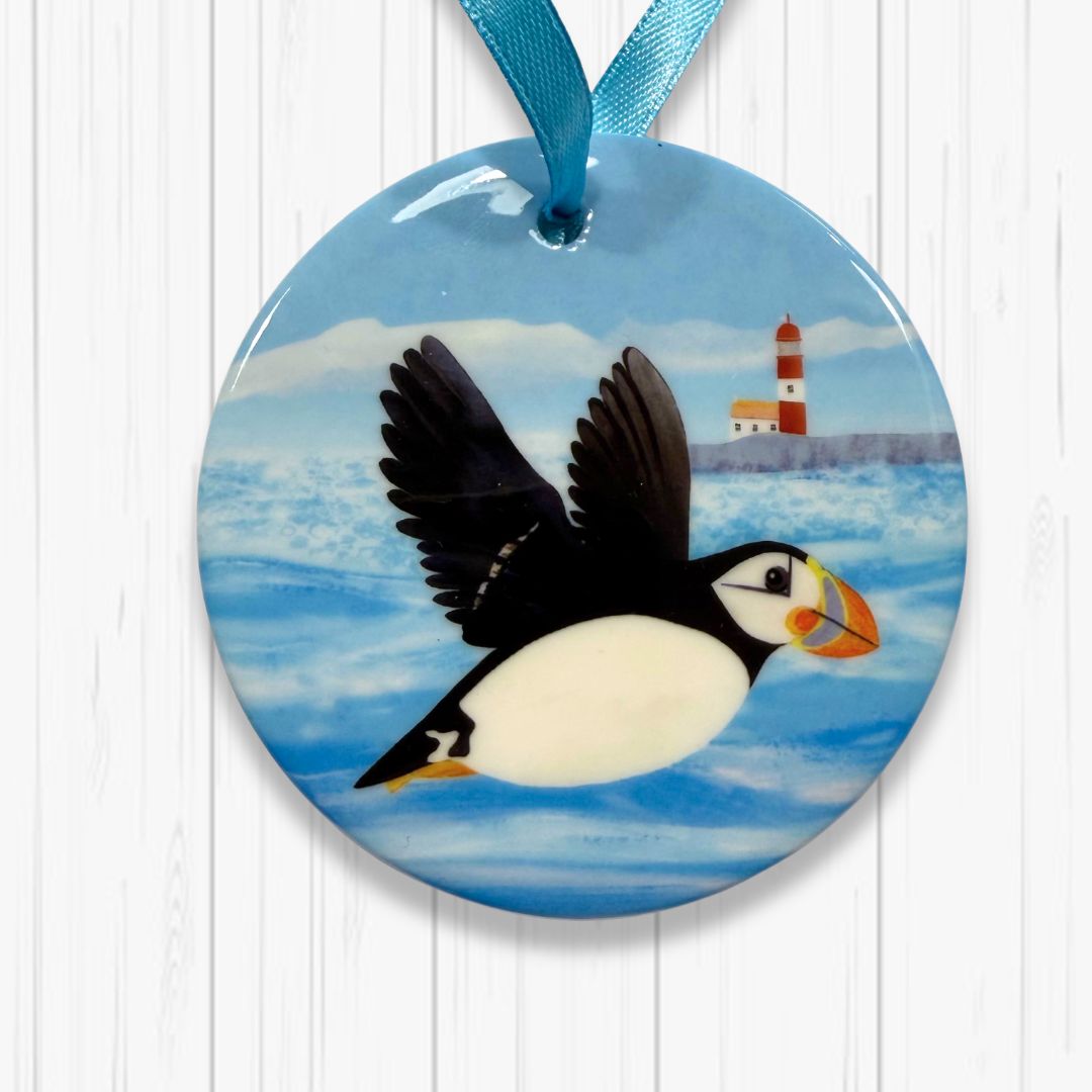 Round ornament with a puffin taking flight and lighthouse design on a blue background.