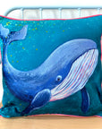 A cushion featuring a blue whale painting on a teal green background with bright pink piping.
