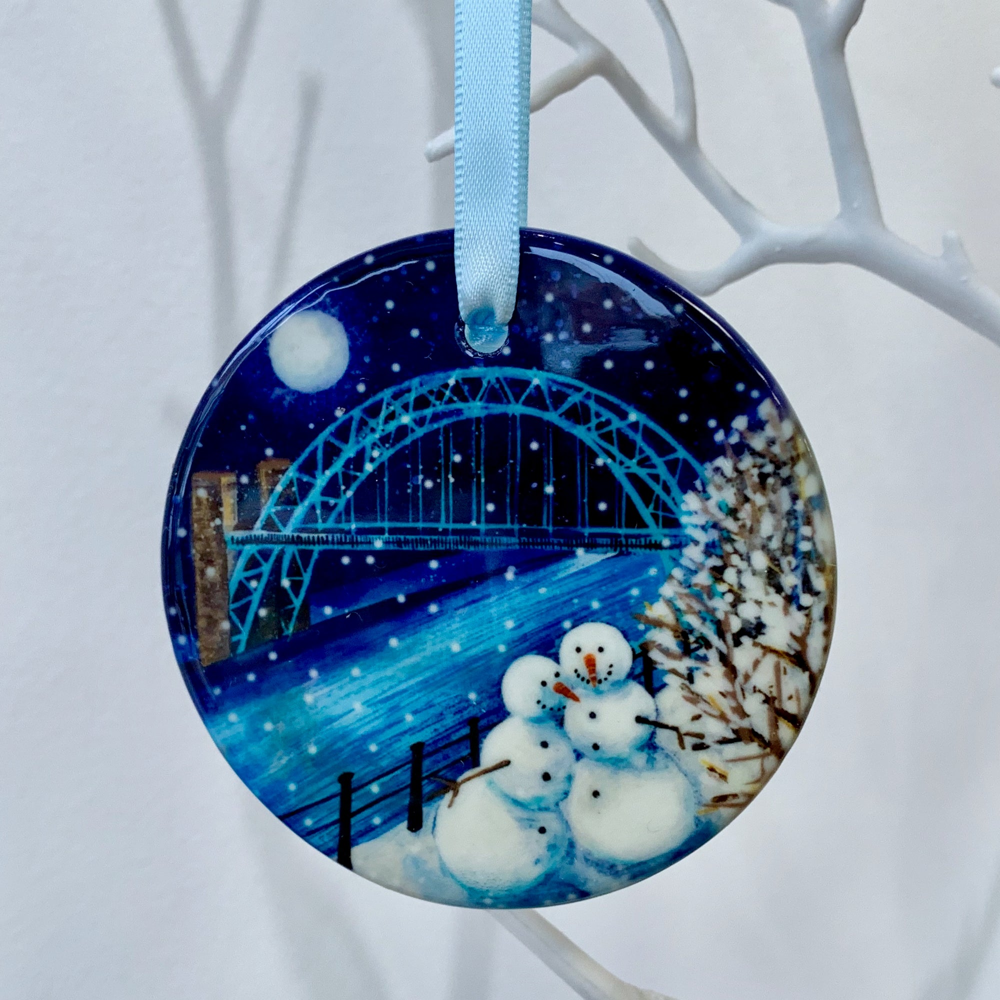 Circular ceramic hanging decoration on a white tree branch. Two smiling snowmen stand side by side by the river with the Tyne Bridge in the background. The sky is dark with a full moon and snow is falling. 