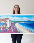 Person holding a large framed artwork of a coastal scene with people and a building labeled 'ICES'.