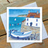 A white building with red roof sits to the right of a calm blue sea with a small sailing boat and rugged coastline beyond. A small boat is depicted in front of the painting and two seagulls fly in the blue cloudless skies.  