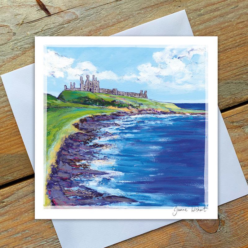 Dunstanburgh castle sits atop a hill along the coastline with a light blue sky, white clouds and rocky shoreline. 