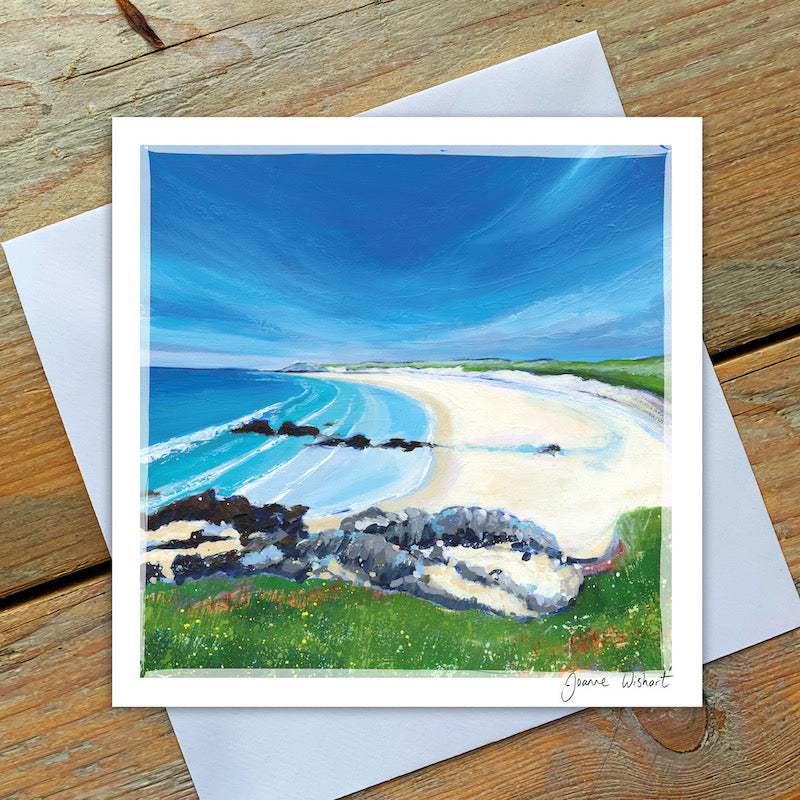 White sands and a calm turquoise sea stretch to the horizon under a deep blue sky with wisps of cloud. A rocky foreground frames the coastal scene.