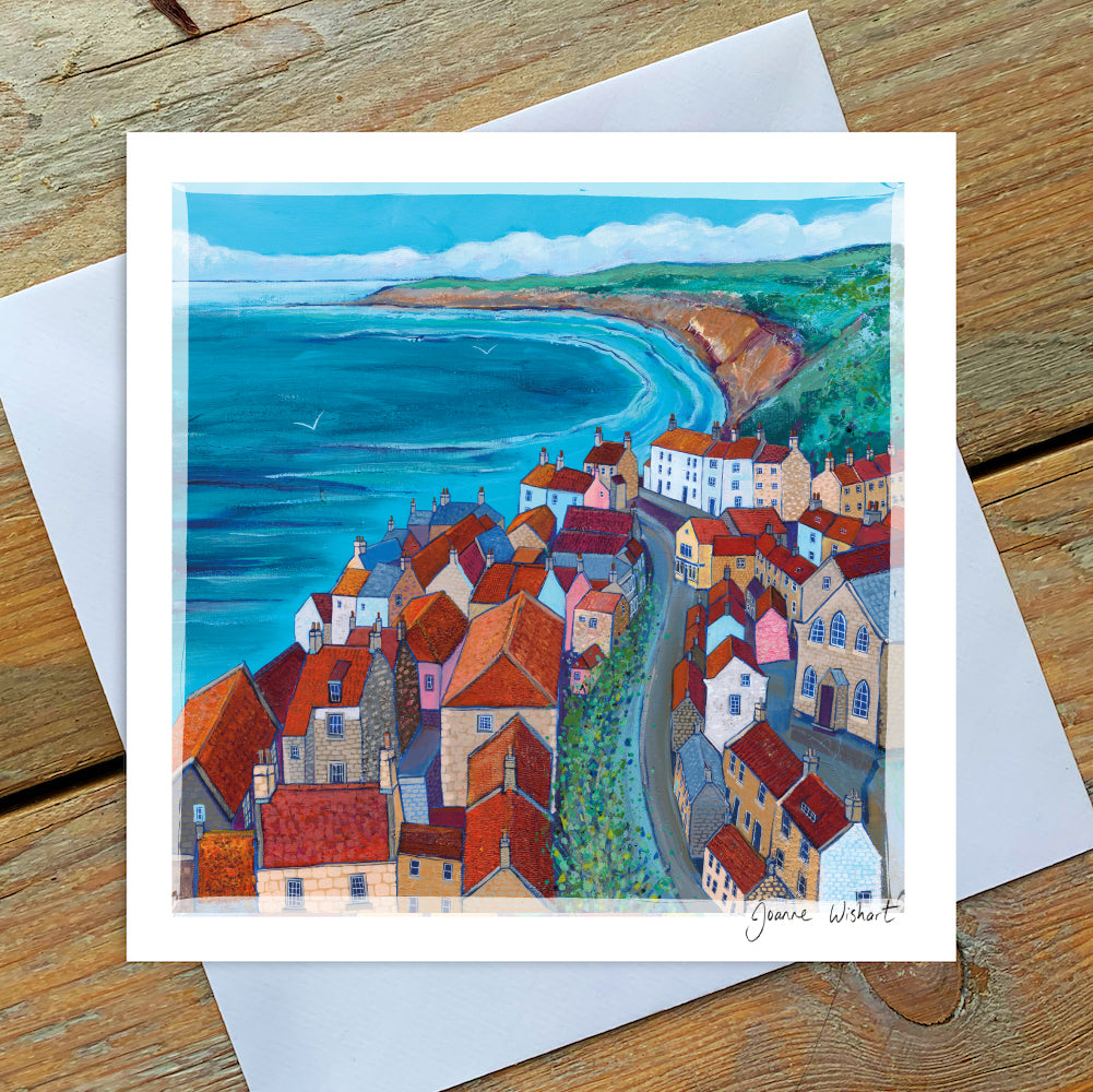 A greetings card of the historic village of Robin Hoods bay showing the orange rooftops with a sea view and cliffs in the distance. 