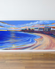 Framed painting of a winter beach scene with buildings and a colourful sky.