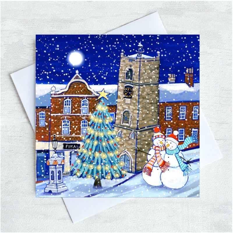 A square Christmas greeting card featuring a snowy Morpeth village scene with a lit Christmas tree, brick buildings, a clock tower, and two snowmen, shown with a white envelope on a light textured background.