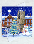 A square Christmas greeting card featuring a snowy Morpeth village scene with a lit Christmas tree, brick buildings, a clock tower, and two snowmen, shown with a white envelope on a light textured background.