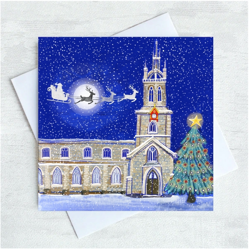 A square Christmas greeting card featuring an illustrated snowy night scene with Newcastle Cathedral in Snow, Santa Claus in a starry blue sky, and a decorated tree, shown with white envelope on a light textured background.
