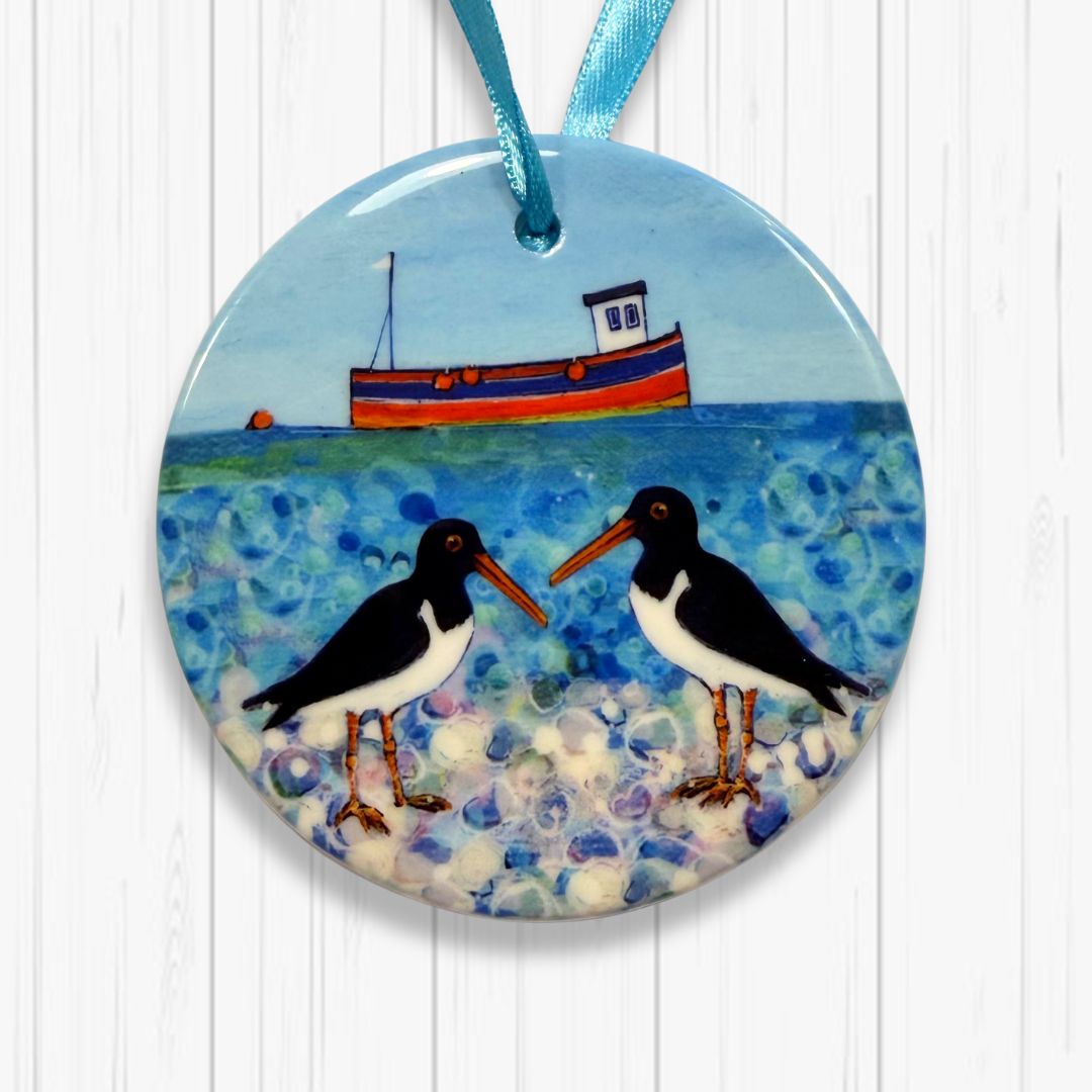 Round ceramic ornament with two Oystercatchers on a beach and a boat in the background, hanging on a white wooden surface.