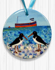 Round ceramic ornament with two Oystercatchers on a beach and a boat in the background, hanging on a white wooden surface.