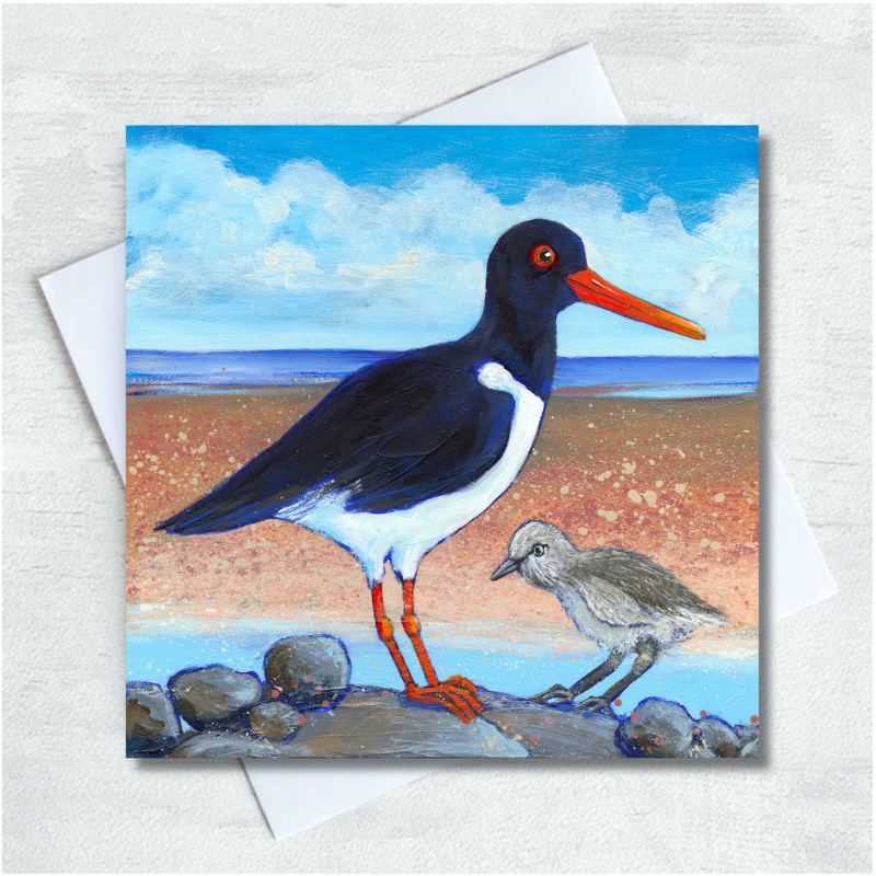 An Oystercatcher and its chick sit atop a grey stony foreground with a sandy beach, blue sea and turquoise cloudy sky behind. 