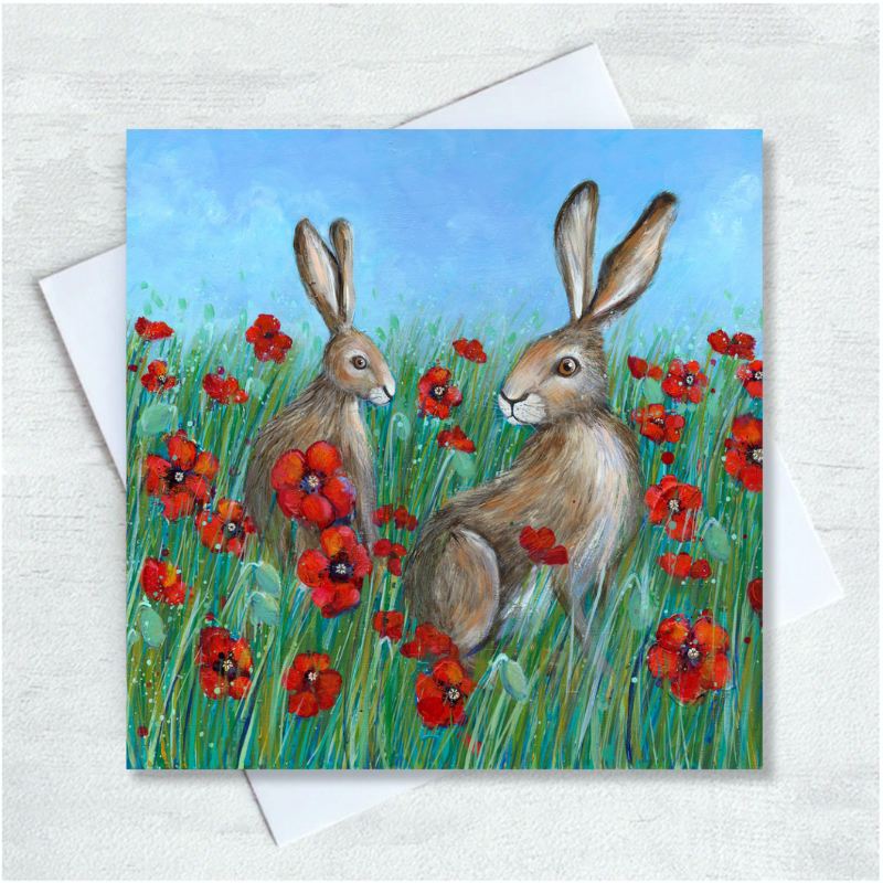 Two brown hairs sit facing inwards amongst a field of long green grass and red poppies. The sky is light blue with a hint of clouds. 