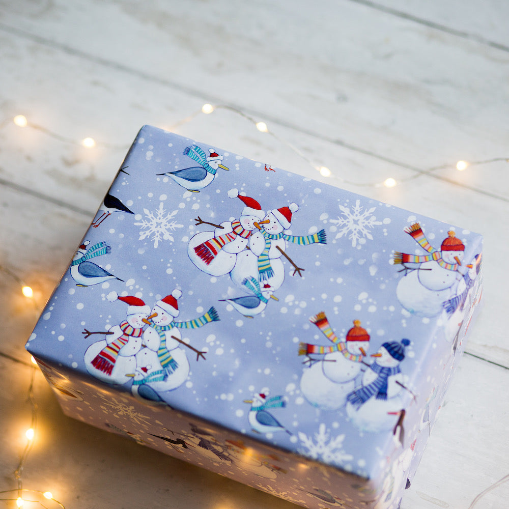 Gift box wrapped in blue paper with snowmen and birds on a wooden surface with fairy lights.