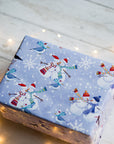 Gift box wrapped in blue paper with snowmen and birds on a wooden surface with fairy lights.