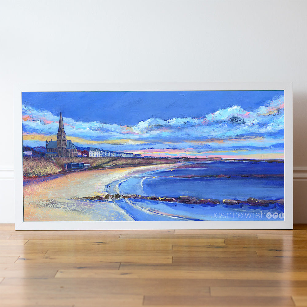 Framed artwork of a coastal scene with a church and beach on a white wall.