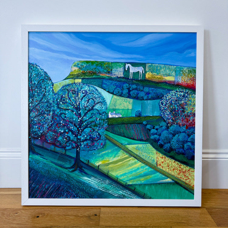 A framed landscape painting leaning against a white wall on a wooden floor. The artwork depicts stylized blue hills, trees, fields, and a white horse on a hilltop under a bright blue sky.