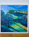A framed landscape painting leaning against a white wall on a wooden floor. The artwork depicts stylized blue hills, trees, fields, and a white horse on a hilltop under a bright blue sky.