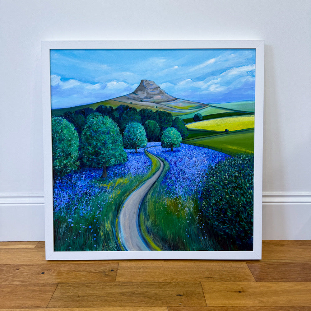 A framed landscape painting of a winding path through a field of blue flowers, with green trees and a mountain under a blue sky, leaning against a white wall on a wooden floor.