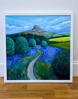 A framed landscape painting of a winding path through a field of blue flowers, with green trees and a mountain under a blue sky, leaning against a white wall on a wooden floor.