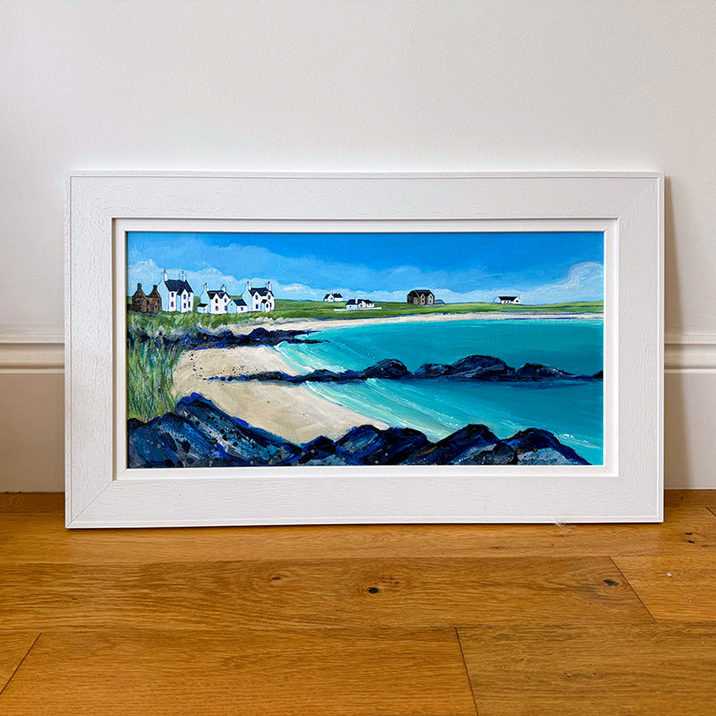 A framed coastal landscape painting leaning against a white wall on a wooden floor. The artwork shows a sandy beach, turquoise water, dark rocky shoreline, green fields, and small white houses under a blue sky, all enclosed in a wide white frame.