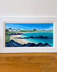 A framed coastal landscape painting leaning against a white wall on a wooden floor. The artwork shows a sandy beach, turquoise water, dark rocky shoreline, green fields, and small white houses under a blue sky, all enclosed in a wide white frame.