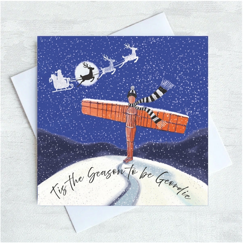 A square Christmas greeting card with a white envelope, featuring an illustration of the Angel of the North and a black-and-white striped scarf standing on a snowy hill under a dark blue night sky with Santa and reindeer flying overhead. The card reads, 'Tis the Season to be Geordie'.
