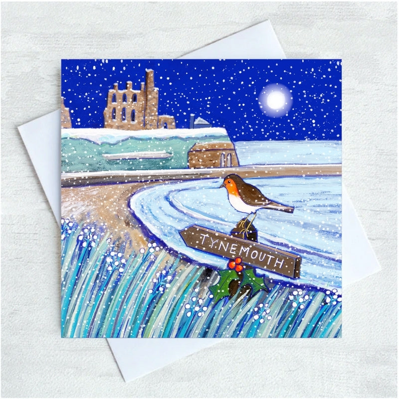 A square illustrated greeting card with a white envelope behind it, showing a snowy coastal scene with Tynemouth Priory in the background with a Robin perched on a sign reading TYNEMOUTH. The card is photographed on a light textured surface.