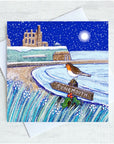 A square illustrated greeting card with a white envelope behind it, showing a snowy coastal scene with Tynemouth Priory in the background with a Robin perched on a sign reading TYNEMOUTH. The card is photographed on a light textured surface.