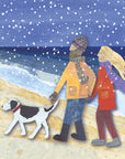 A loving vouple walk their dog on the beach during a snow flurry. They are wearing colourful long scarfs which blow in the wind behind them. 