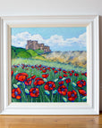 A bold and colourful original painting in a white wood frame. It sets resting ona wood floor against a white wall. The painting is of Bamburgh Castle with bright red poppies in the foreground.
