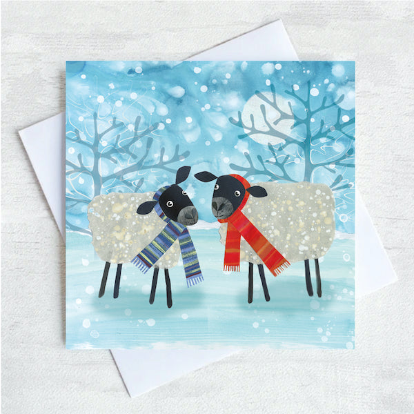 A christmas greetings card featuring two black headed sheep facing one another wearing brightly coloured scarfs. They are standing in a field of show with snowflakes falling all around them. 