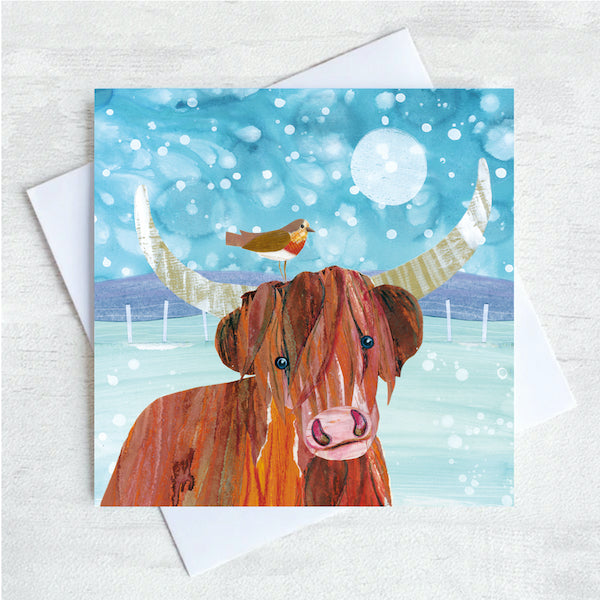 A Christmas card featuring a Highland Cow and a robin perched on one if its horns. THey stand in a fields with snow fluttering overhead. 
