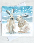 A winter themed greetings card featuring a collage illustration of two winter snow hares in a snow scene.