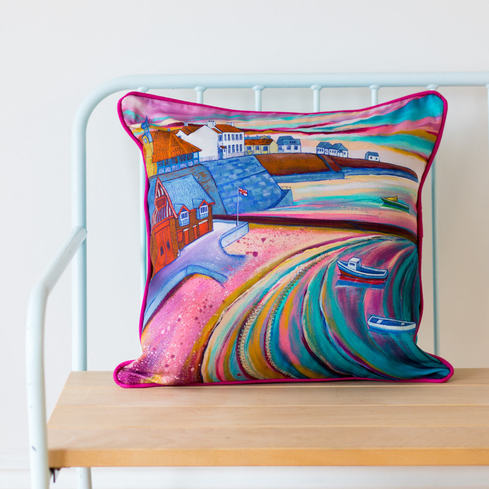 A bright pink cushion featuring a scene of cullercoast harbour sits on a blue chair with a white background. 