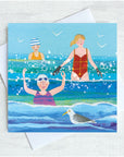 Three ladies and a seagull splash amongst the waves at the seaside. The are wearing broightly coloured swimsuits and have rosy red cheeks. 