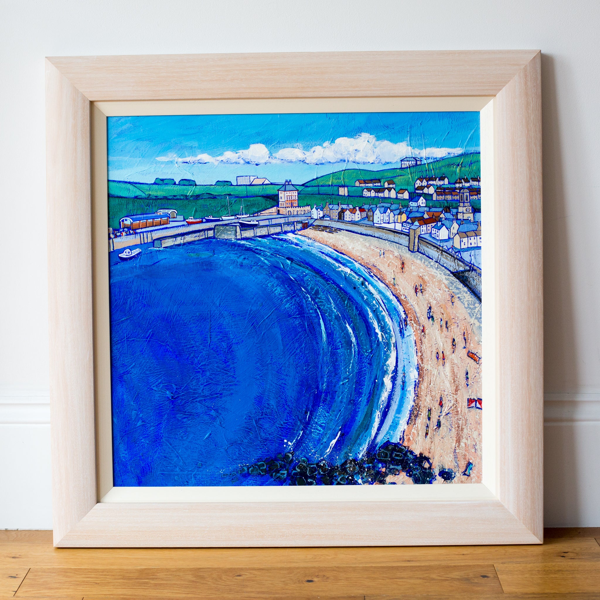 A coastal painting of Eyemouth Harbour in a limewaxed chuncky wood frame. It sits on a wood floor and is leaning against a white wall. 
